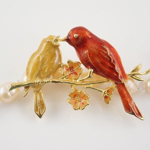 May include: A gold-toned brooch featuring two birds perched on a branch with small flowers. One bird is yellow, the other is red-orange. The birds are detailed with enamel and the branch has a gold finish. The brooch is set against a backdrop of white pearls.