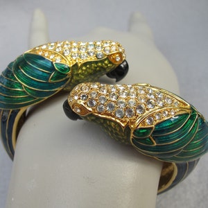 May include: A gold tone bangle bracelet with two parrot figures. The parrots are green and blue with gold accents and are covered in clear rhinestones.