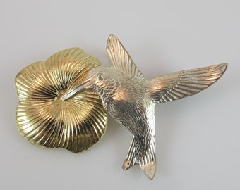 May include: A silver hummingbird brooch with a gold flower design. The hummingbird is in flight with its wings spread out. The flower is a stylized design with a center and petals.
