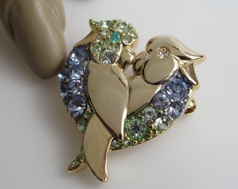 May include: Gold-tone brooch featuring two parrots facing each other, their bodies forming a heart shape. The parrots are adorned with blue, green, and clear rhinestones.