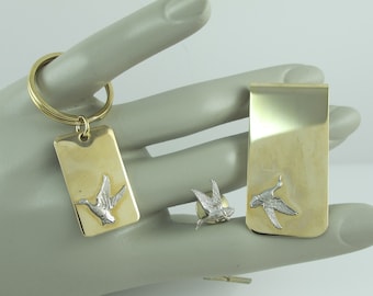 May include: A set of three gold-toned metal accessories featuring a flying duck design. The set includes a keychain, a money clip, and a tie tack.