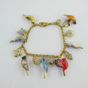 May include: A gold-toned charm bracelet featuring colorful bird charms, flowers, and other nature-inspired elements. The bracelet includes a variety of bird designs in yellow, blue, red, and orange, along with a sunflower, acorn, and leaf charms. The bracelet is approximately 7 inches long.