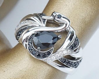 May include: A silver ring with a pear-shaped black gemstone. The ring has a snake design with black and clear accent stones. The ring is displayed on a gold-toned surface.
