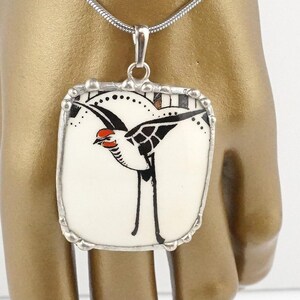 May include: A silver pendant necklace featuring a square, cream-colored pendant with a black and red bird design. The bird is depicted in a stylized, art deco style. The pendant is framed with a silver border and hangs from a delicate silver chain.