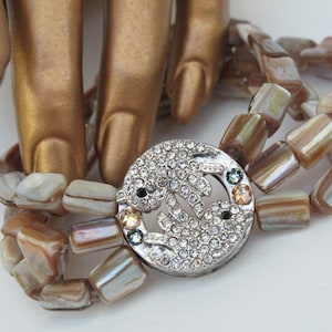 May include: A multi-strand bracelet made of iridescent brown and white shell beads with a silver-toned clasp featuring a design of two fish encrusted with clear rhinestones.