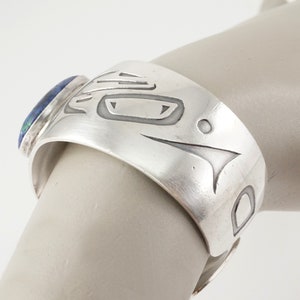 May include: Silver cuff bracelet with a blue and green stone set into the side. The bracelet is decorated with a carved design of a stylized animal face.