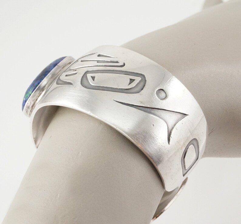 May include: Silver cuff bracelet with a blue and green stone set into the side. The bracelet is decorated with a carved design of a stylized animal face.