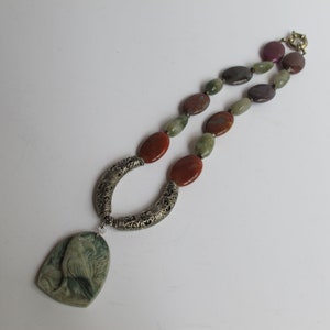 May include: A silver necklace with a carved bird pendant and multi-colored agate beads. The beads are in shades of green, brown, and purple.
