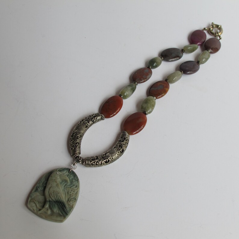 May include: A silver necklace with a carved bird pendant and multi-colored agate beads. The beads are in shades of green, brown, and purple.