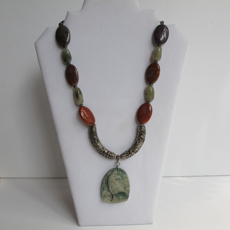 May include: A multi-colored stone necklace with a silver pendant featuring a carved bird design. The necklace features brown, green, and purple stones.
