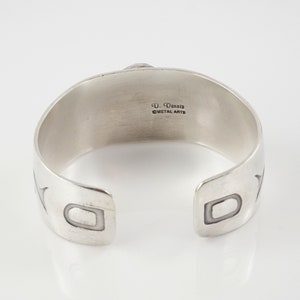 May include: Silver cuff bracelet with a geometric design and the inscription "D. Dennis Metal Arts" on the back.