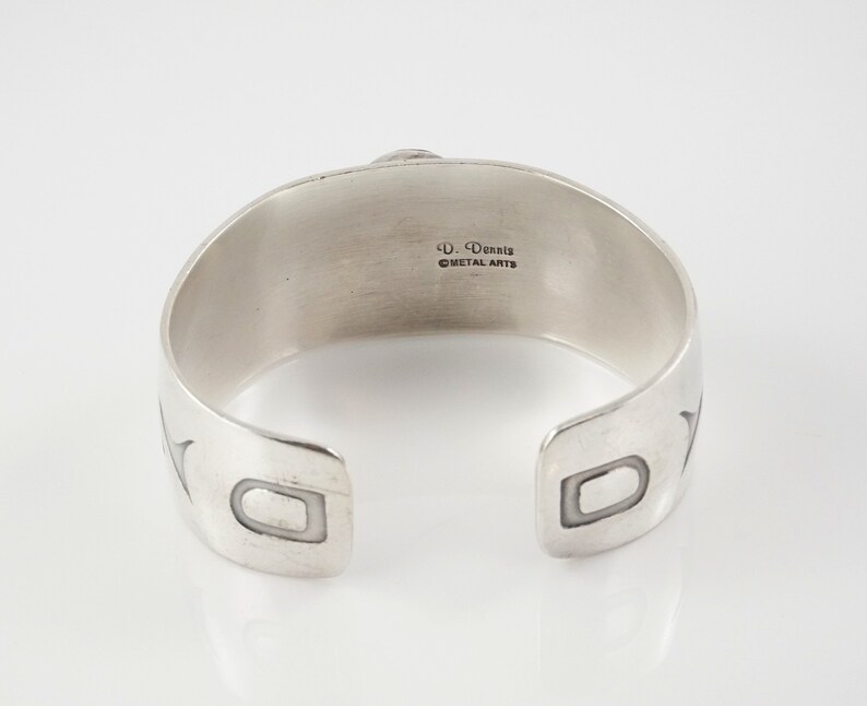 May include: Silver cuff bracelet with a geometric design and the inscription "D. Dennis Metal Arts" on the back.