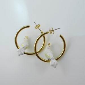 May include: A pair of gold-colored hoop earrings, each featuring a small, white bird with yellow and gold accents. The birds are perched on the hoops, creating a unique and whimsical design. The earrings are set against a white background.