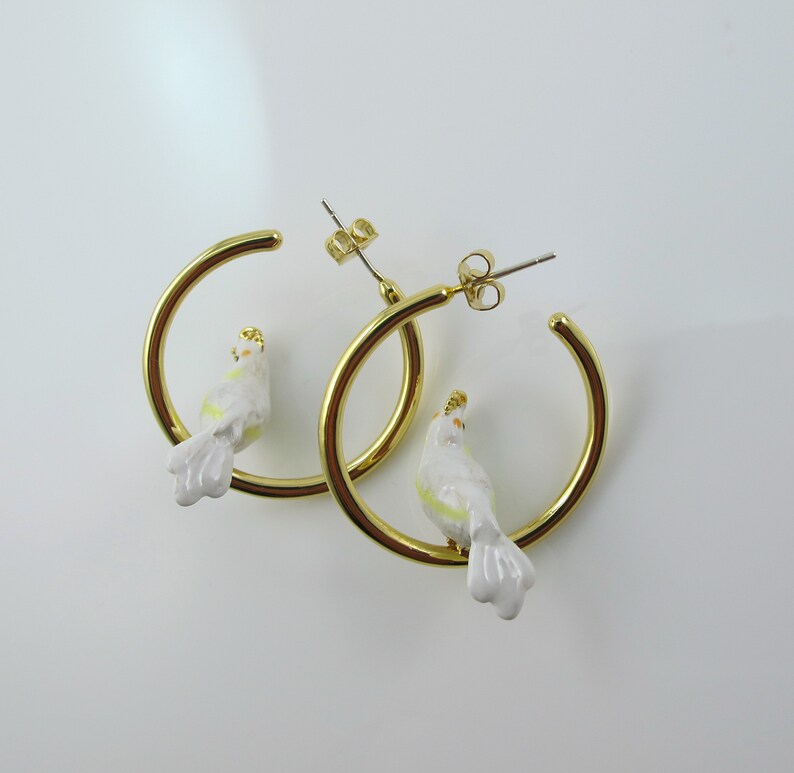 May include: A pair of gold-colored hoop earrings, each featuring a small, white bird with yellow and gold accents. The birds are perched on the hoops, creating a unique and whimsical design. The earrings are set against a white background.
