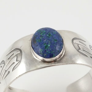 May include: A silver bracelet with a blue and green gemstone set in the center. The bracelet has a design of stylized birds on either side of the stone.