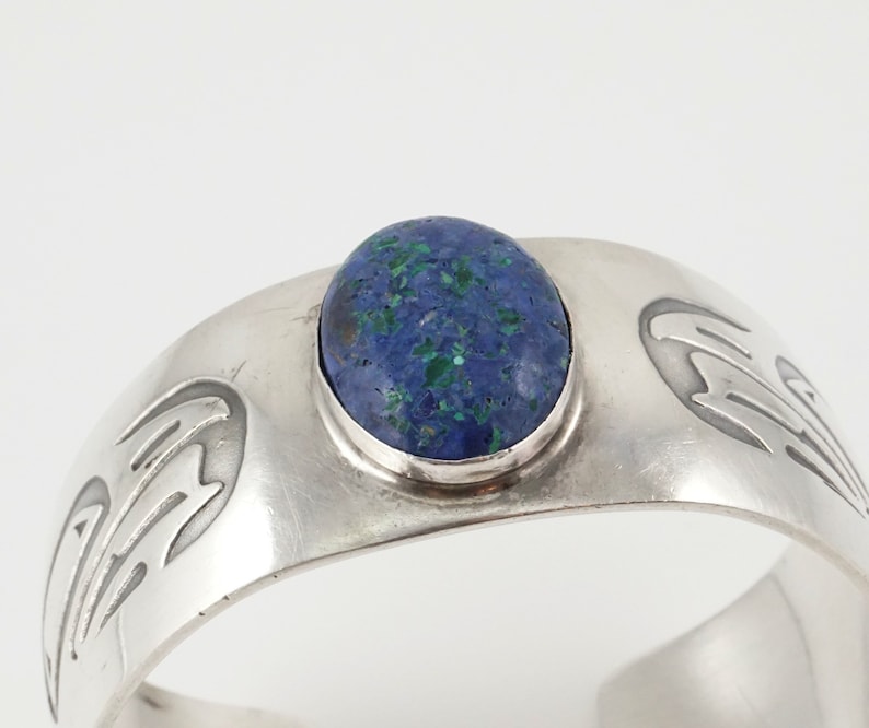 May include: A silver bracelet with a blue and green gemstone set in the center. The bracelet has a design of stylized birds on either side of the stone.