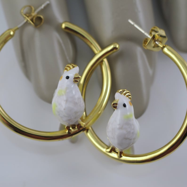 May include: A pair of gold-colored hoop earrings. Each earring features a small, white bird with yellow and gold accents perched on the hoop. The earrings are displayed against a neutral background.