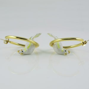 May include: A pair of gold-colored hoop earrings, each featuring a small, white bird with yellow and green accents. The birds appear to be perched on the hoops, with their beaks and feet visible. The earrings are set against a white background.