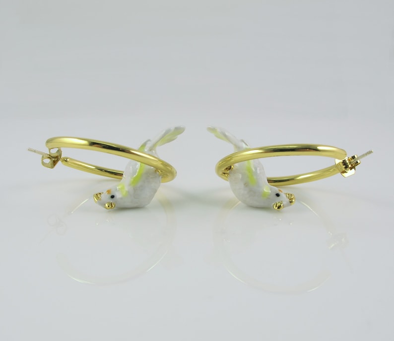 May include: A pair of gold-colored hoop earrings, each featuring a small, white bird with yellow and green accents. The birds appear to be perched on the hoops, with their beaks and feet visible. The earrings are set against a white background.