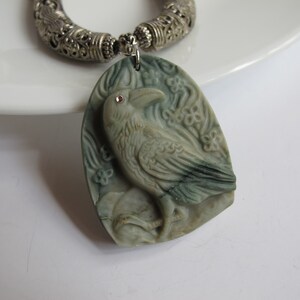 May include: A green stone pendant necklace with a carved raven design. The raven has a small red rhinestone for an eye. The necklace has a silver chain with a decorative silver clasp.