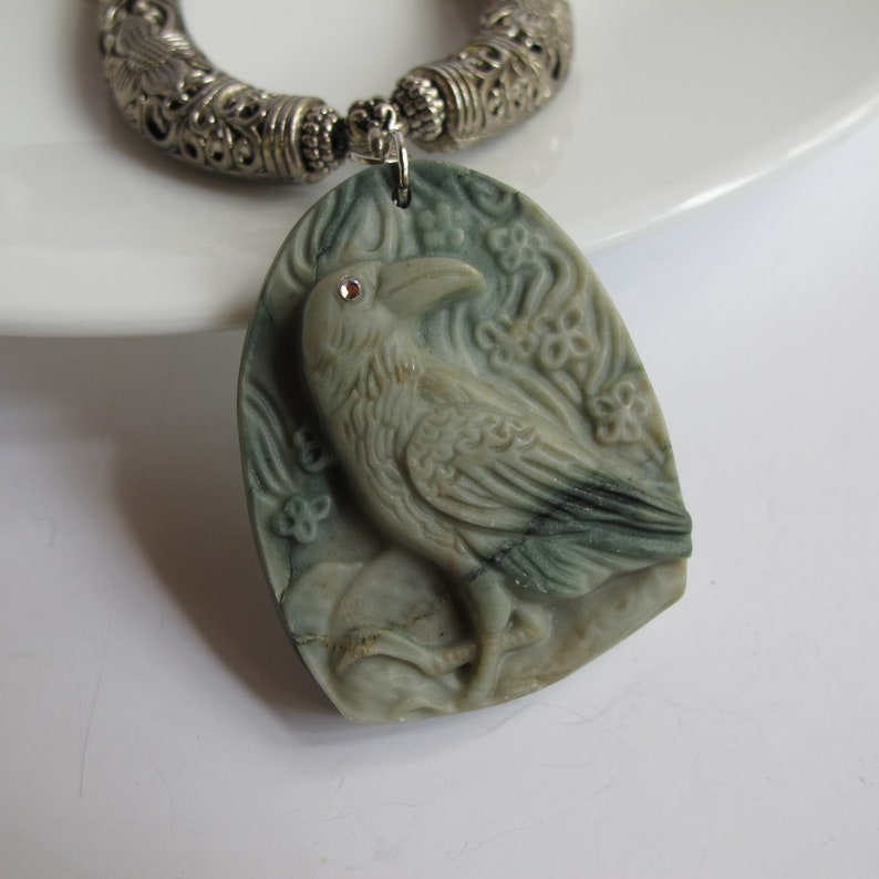May include: A green stone pendant necklace with a carved raven design. The raven has a small red rhinestone for an eye. The necklace has a silver chain with a decorative silver clasp.