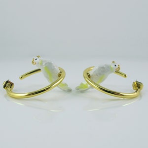 May include: A pair of gold-colored hoop earrings, each featuring a white and yellow bird design. The birds are perched on the hoops, with their heads and tails extending outwards. The earrings have a post-back closure.