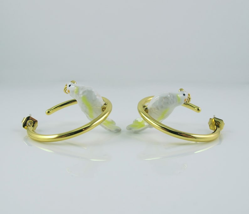 May include: A pair of gold-colored hoop earrings, each featuring a white and yellow bird design. The birds are perched on the hoops, with their heads and tails extending outwards. The earrings have a post-back closure.