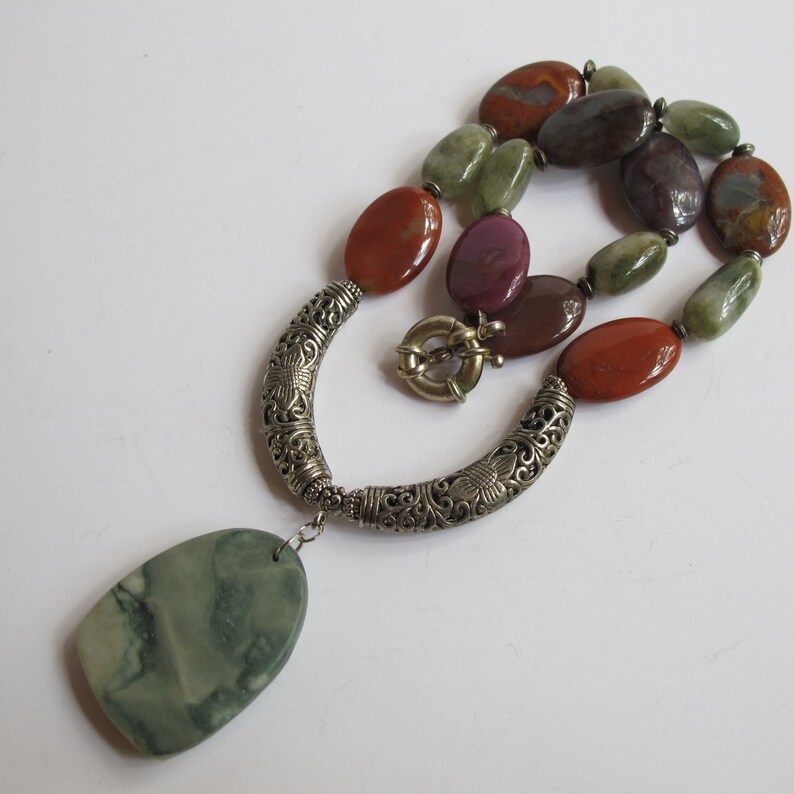 May include: A silver necklace with a large green stone pendant and a silver clasp. The necklace is made with a variety of colorful stones, including red, green, and purple.