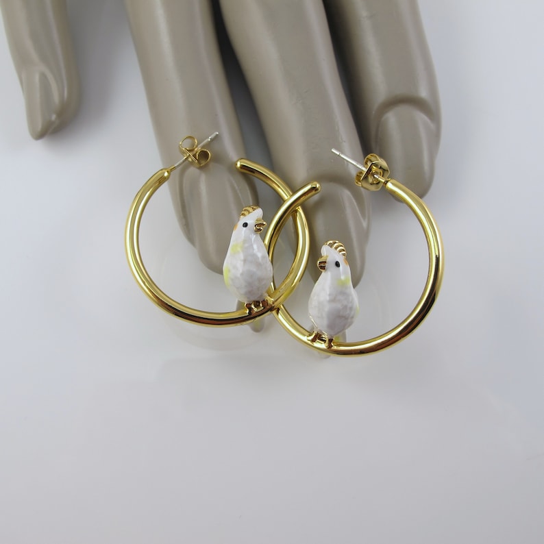 May include: Gold-tone hoop earrings featuring a white and yellow bird charm. The earrings have a polished finish and a post back closure. The bird charms have a detailed design, adding a unique touch to the jewelry.