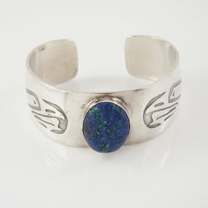 May include: A silver cuff bracelet with a blue oval gemstone set in the center. The bracelet has a stylized design on either side of the stone.
