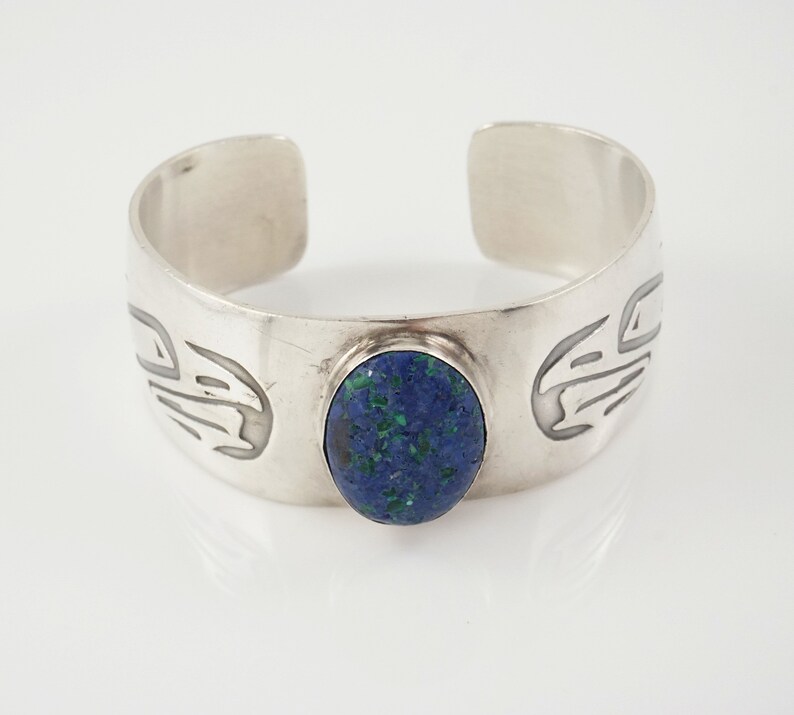 May include: A silver cuff bracelet with a blue oval gemstone set in the center. The bracelet has a stylized design on either side of the stone.