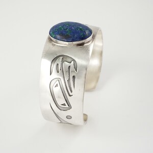 May include: A silver cuff bracelet with a blue and green stone set in the center. The bracelet is decorated with a carved design.