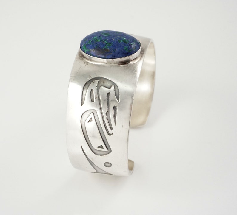 May include: A silver cuff bracelet with a blue and green stone set in the center. The bracelet is decorated with a carved design.