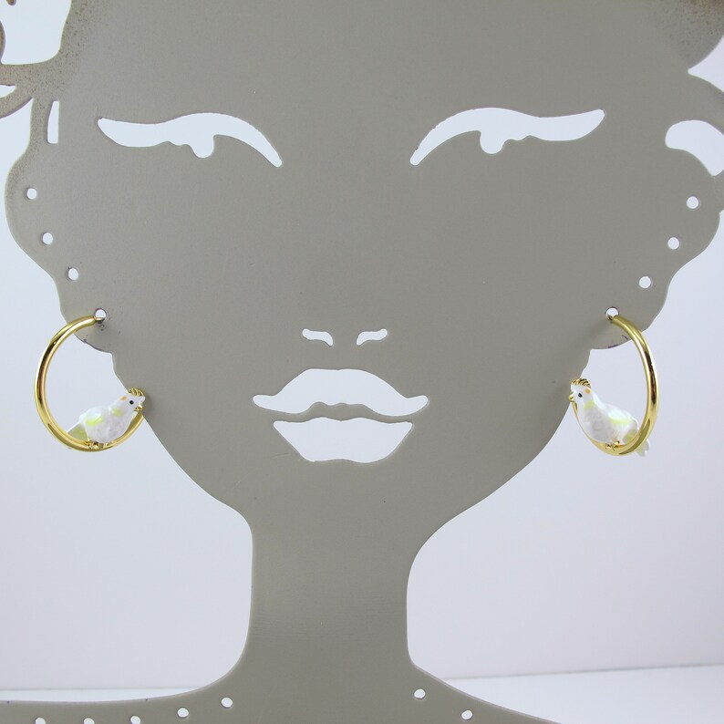 May include: Gold-tone hoop earrings featuring a white bird design with iridescent accents. The bird has a yellow crown. The earrings are displayed on a gray mannequin head.