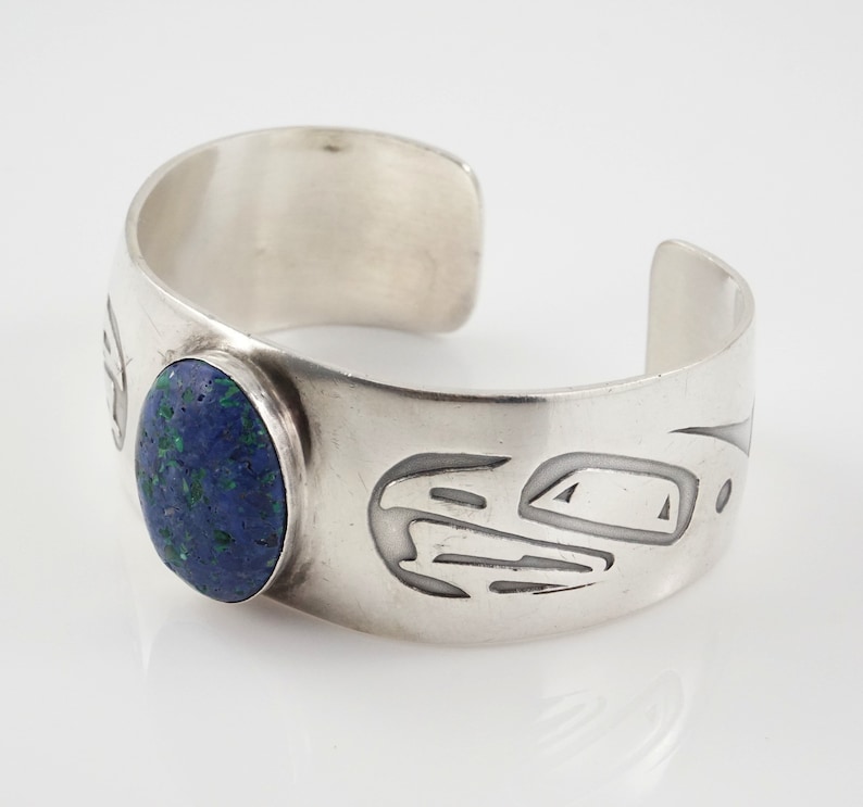 May include: A silver cuff bracelet with a blue and green stone set in the center. The bracelet is decorated with a carved design.