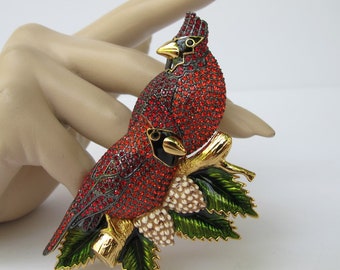 May include: A decorative brooch featuring two red cardinal birds perched on a gold-colored branch with green leaves and white berries. The birds are covered in red rhinestones with black accents. The brooch is a detailed piece of jewelry.
