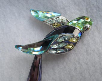 May include: A crystal bird figurine with iridescent wings and a green head. The bird has a silver metal body and is adorned with various colored crystals. The tail is a deep purple color. The bird is a decorative item.