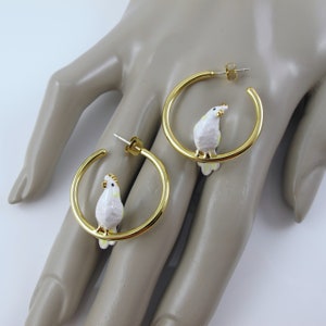 May include: A pair of gold-colored hoop earrings, each featuring a white and yellow parrot charm. The parrot charms are perched on the hoops. The earrings are displayed on a light-colored surface.
