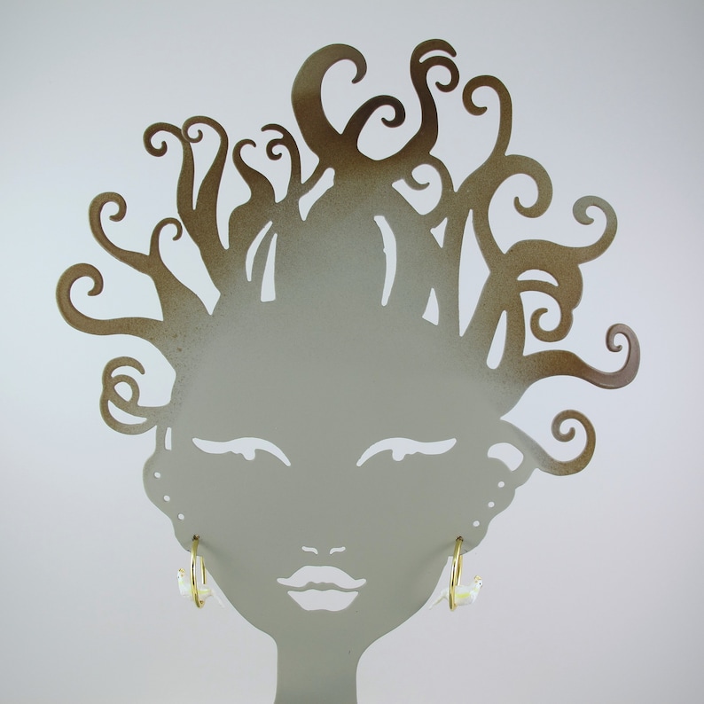 May include: A gold-colored metal earring display stand shaped like a woman's face with curly hair. Two gold hoop earrings with white and yellow accents are displayed on the stand. The background is plain white.