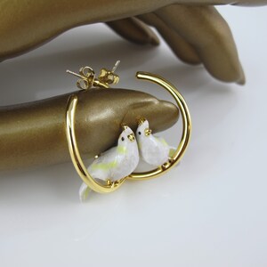 May include: Gold-tone hoop earrings featuring two white and yellow bird figures. The birds are perched on the hoop, adding a whimsical touch to the jewelry. The earrings have a post back closure.