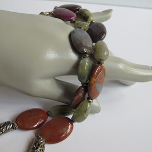 May include: A multi-strand necklace with large, oval-shaped gemstone beads in shades of red, green, and brown. The beads are separated by small, silver-toned spacers.