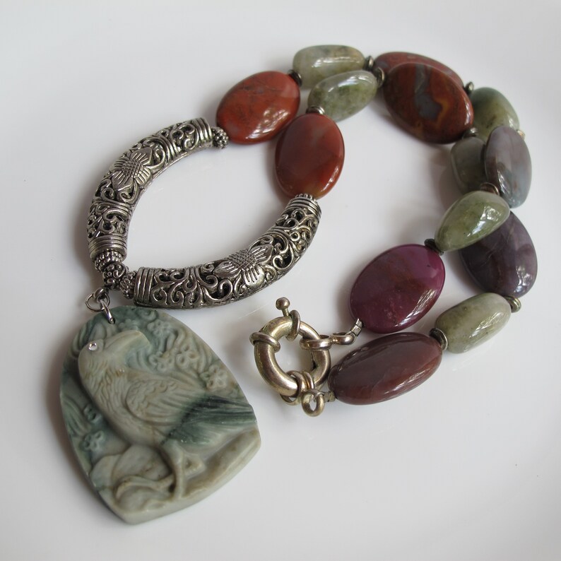 May include: A silver necklace with a carved stone bird pendant. The necklace features a silver chain with a decorative clasp and is adorned with multi-colored agate beads.