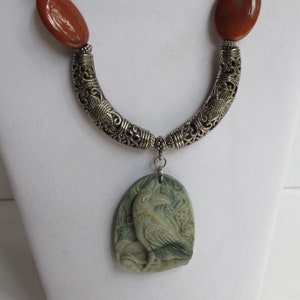 May include: A silver necklace with a carved green stone pendant featuring a detailed raven design. The pendant has a small red rhinestone for an eye. The necklace has two brown stone beads at the top.