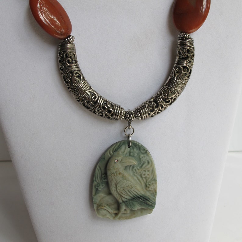 May include: A silver necklace with a carved green stone pendant featuring a detailed raven design. The pendant has a small red rhinestone for an eye. The necklace has two brown stone beads at the top.