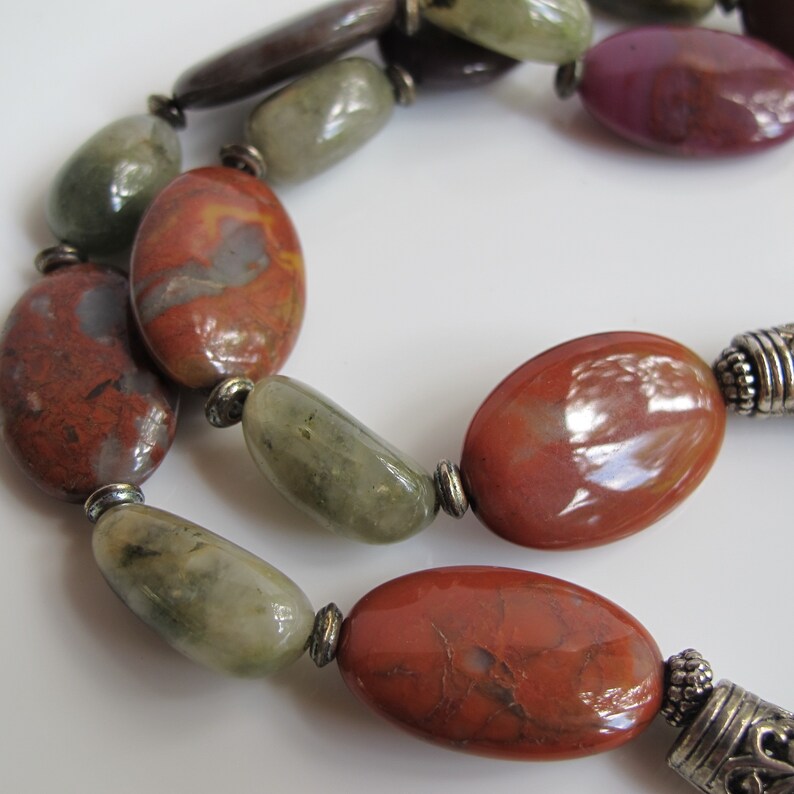 May include: A close-up of a necklace made with red, green, and brown oval-shaped stones. The stones are separated by silver metal beads.