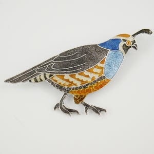 Op de afbeelding: Een broche van zilver en emaille in de vorm van een kwartel. De kwartel is afgebeeld in een realistische stijl met bruin, oranje, wit en blauw emaille.