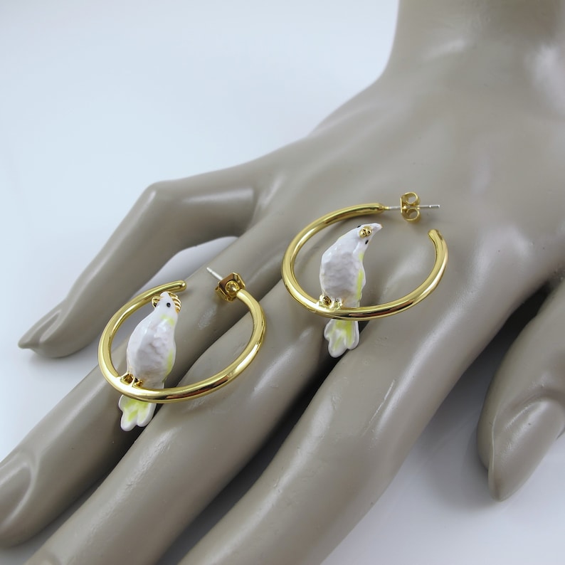 May include: Gold-tone hoop earrings featuring a white and yellow parrot design. The parrot is perched on the hoop, adding a whimsical touch. These earrings are a unique accessory for those who love bird-themed jewelry.