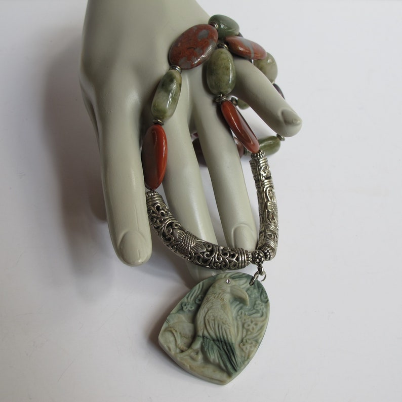 May include: A silver necklace with a carved green stone raven pendant and a multi-colored stone beaded chain.
