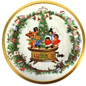 Puede incluir: Una bandeja redonda de metal con borde dorado que presenta una ilustración central de niños con diversas vestimentas tradicionales bailando alrededor de un árbol de Navidad decorado sobre una caja de música, rodeado por una corona de ramas de pino y banderas.
