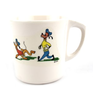 Puede incluir: Taza de cerámica blanca con los personajes de dibujos animados Pluto y Goofy corriendo. Pluto lleva un atuendo rojo, y Goofy viste una camisa amarilla y pantalones azules. La taza tiene un asa curva.